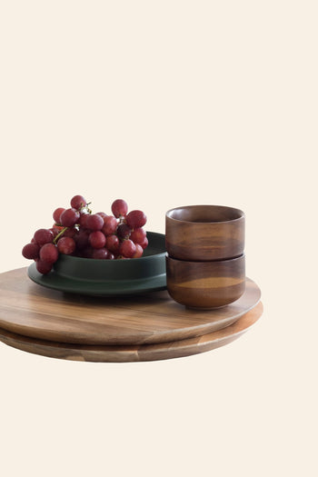 A Serax Collage Tea Cup in brown rests on a green plate beside two stacked acacia wood bowls, all arranged on round wooden boards against a plain light background.