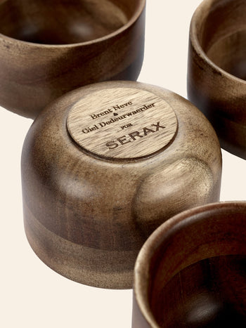A close-up of four round Serax Collage Tea Cups in brown acacia wood, one upside down showing Brent Neve, Giel Dedeurwaerder for SERAX engraved on the base, from the collection inspired by Japanese tea cups.