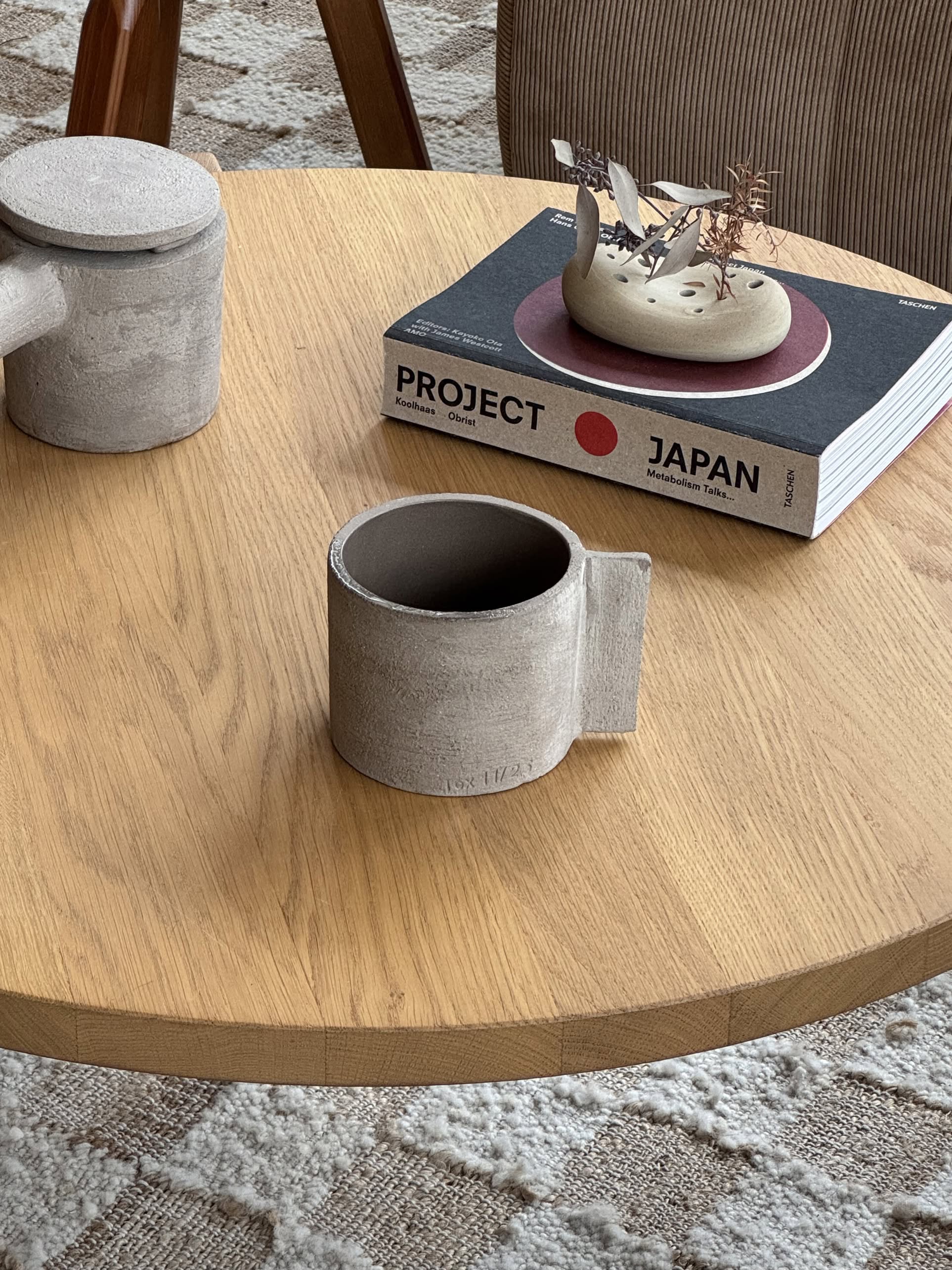 A round wooden table features the Serax Fck Cup L - Grey Brown, a matching teapot, the black and white book PROJECT JAPAN, and a small decor piece on a patterned rug, channeling brutalist minimalism.