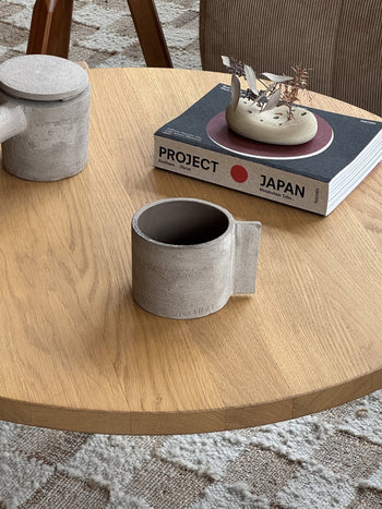 A round wooden table features the Serax Fck Cup L - Grey Brown, a matching teapot, the black and white book PROJECT JAPAN, and a small decor piece on a patterned rug, channeling brutalist minimalism.