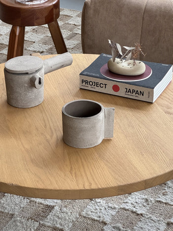 A wooden table with a gray ceramic teapot and the Serax Fck Cup L - Grey Brown, a stack of books topped with a minimalist sculpture in concrete style, and a striped chair in the background. The top book is PROJECT JAPAN.