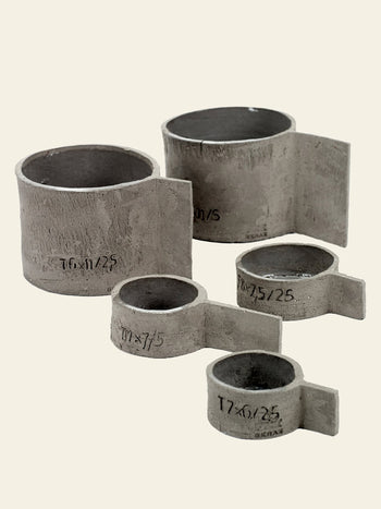 Five Serax Fck Cup L - Grey Brown vessels, featuring a refined concrete look and flat extensions, are arranged on a light beige background. Each cup by Serax has stamped text to indicate its size and type, inspired by brutalist design.