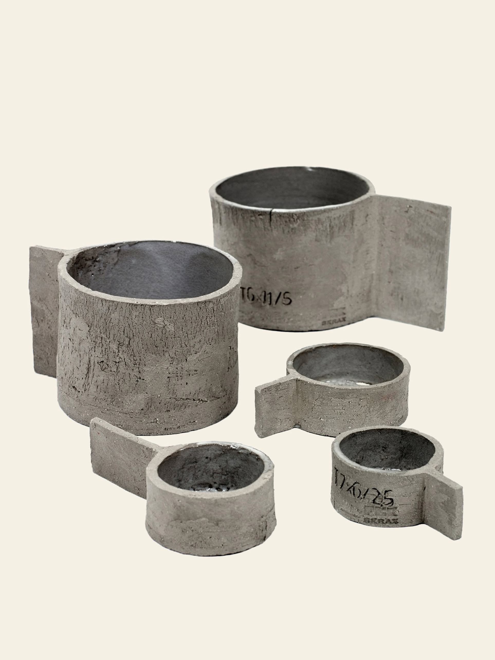 Five Serax Fck Cup L - Grey Brown pieces, each cylindrical with a flat handle-like extension, showcase Serax’s concrete-inspired design reminiscent of brutalist architecture, arranged on a plain, light background.