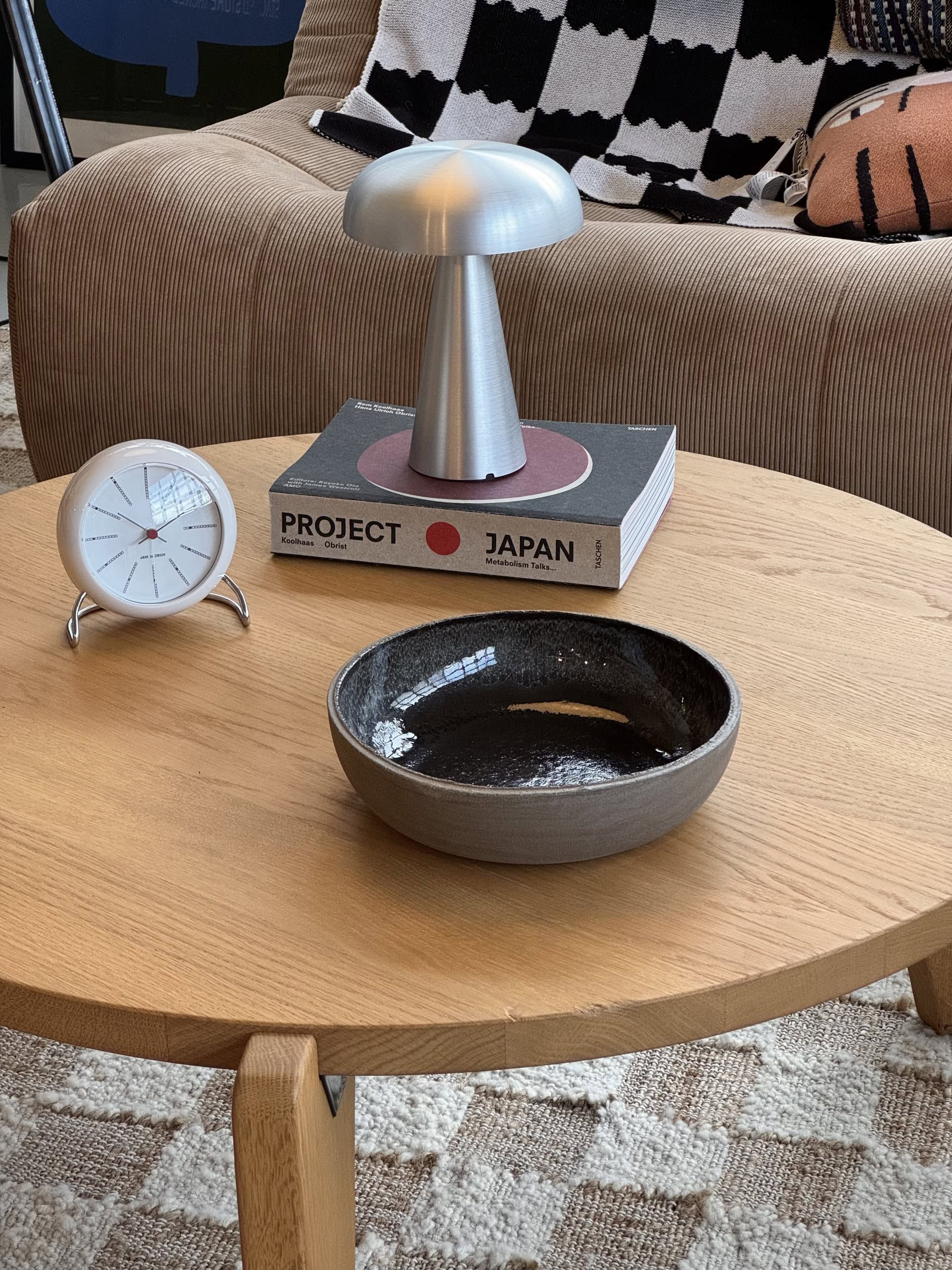A round wooden coffee table holds a Serax Fck Deep Plate in grey, a modern silver desk clock, and two stacked books topped with a mushroom-shaped silver lamp. In the background is a beige sofa with patterned textiles.