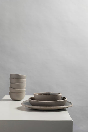 A stack of four small beige ceramic bowls sits beside Serax Fck Deep Plates and bowls in grey, their minimalist, neutral tones inspired by brutalist architecture, all arranged on a white surface against a plain gray background.