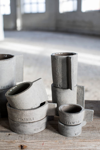 A collection of Serax Fck Teapot N°3 - Grey Brown ceramics with handles and stamped numbers is displayed on a wooden surface in a sunlit, industrial-style room with large windows.