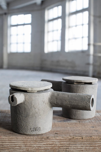 Two grey-brown Serax Fck Teapot N°3 by Serax with side spouts and flat lids are placed on a wooden surface in a spacious, bright room with tall windows and a concrete floor.