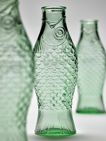 Three Serax Fish & Fish Carafes in green, shaped like textured fish with detailed scales and fins, stand on a plain light background. The central Serax bottle is in sharp focus while the others appear softly blurred behind.