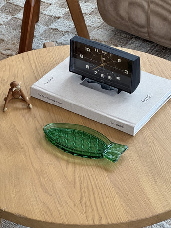 A round wooden table holds the Serax Fish & Fish Serving Dish N°1 - Green by Serax, a wooden figurine, the book The Art of Creating a Home, and a black vintage-style clock.