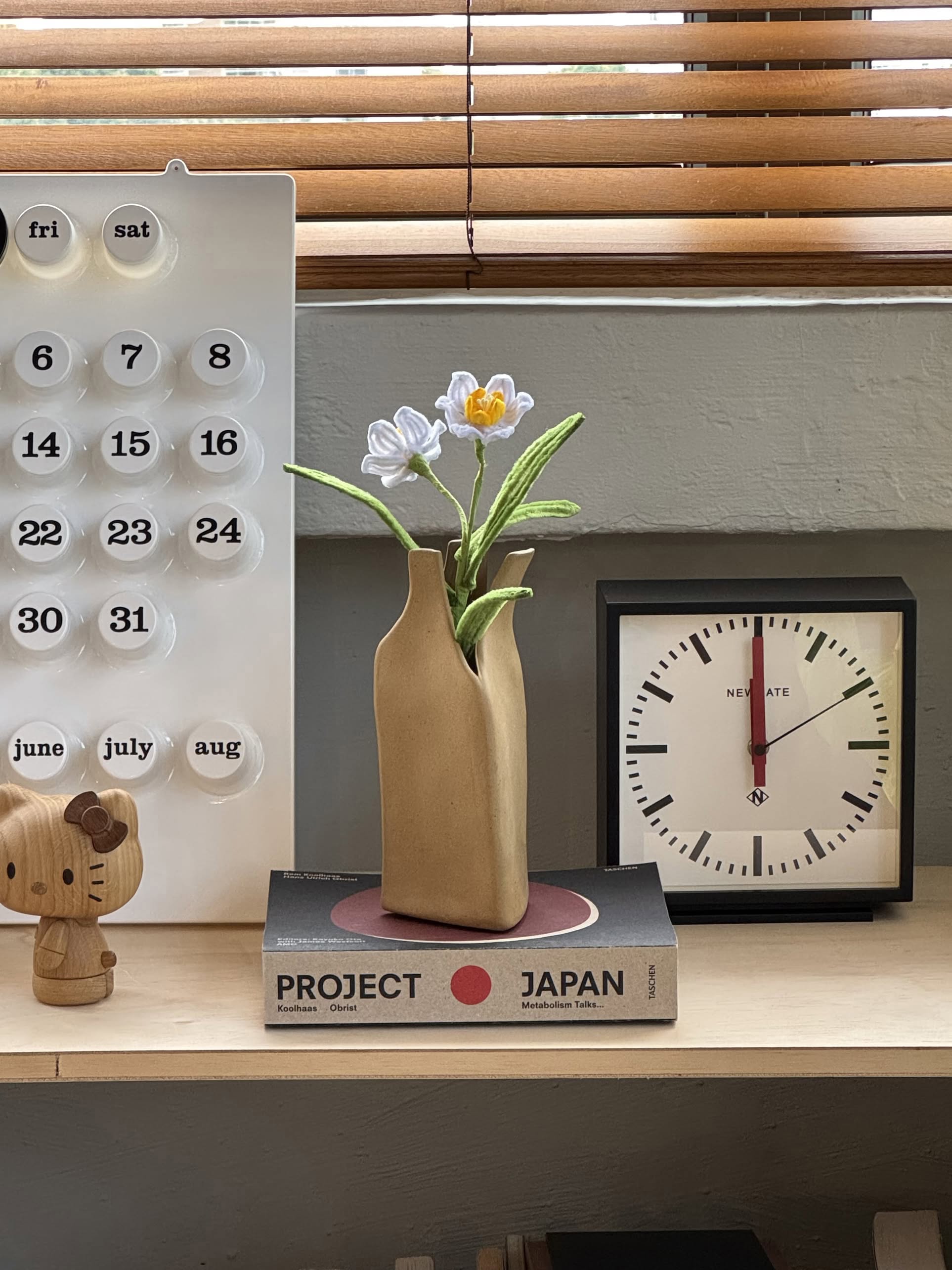 A wooden shelf with a black clock at 12:00, a calendar, a wooden bear figurine, two stacked books titled PROJECT and JAPAN, and the Serax Flower Bottles Vase M - Brown by Serax with white flowers—ideal for minimalist home decor.