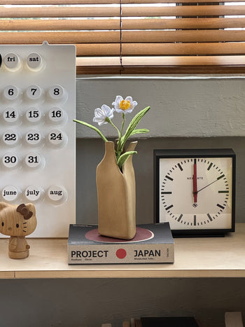 A wooden shelf with a black clock at 12:00, a calendar, a wooden bear figurine, two stacked books titled PROJECT and JAPAN, and the Serax Flower Bottles Vase M - Brown by Serax with white flowers—ideal for minimalist home decor.