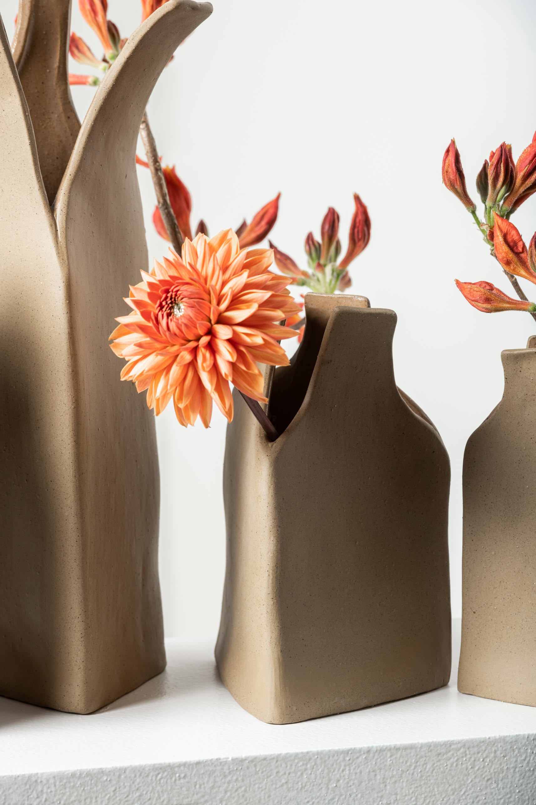 Three Serax Flower Bottles Vase M - Brown by Serax, with a matte tan ceramic finish, hold orange flowers—highlighted by a bold dahlia—set against a white background for modern, minimalist elegance.