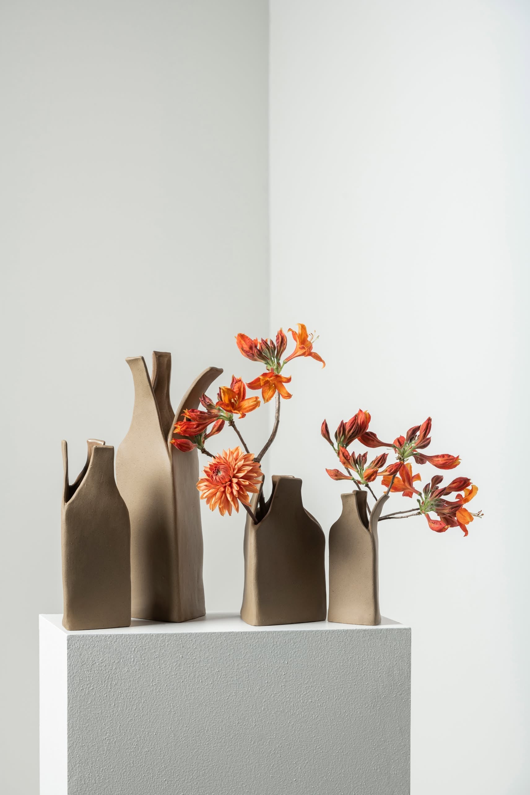 Two Serax Flower Bottles Vase XS - Brown by Serax, containing orange and red flowers, and a third matte brown vase with geometric shapes by Nadia Gallardo, are displayed on a white pedestal for a vibrant minimalist look.