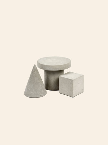 Three geometric shapes—a cone, a cylinder with a flat circular top, and the Serax Marie Cube Concrete - Grey by Serax—are arranged on a light beige background, lending an industrial vibe to modern decor.