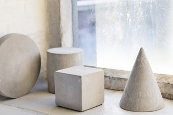Four concrete geometric shapes—a cylinder, a sphere, the Serax Marie Cube Concrete - Grey, and a cone—sit on a windowsill before a frosty window, bringing an industrial accent to modern decor.