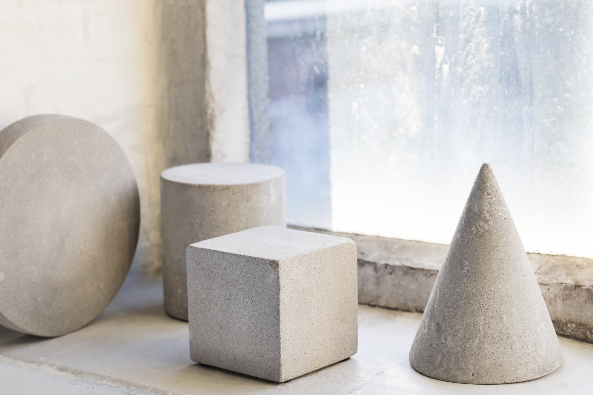 Geometric concrete shapes—a sphere, cube, cone, and Serax Marie Cylinder Concrete - Grey—are arranged on a windowsill near a large window with soft natural light, bringing minimalism and an industrial vibe.