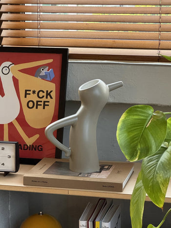 A quirky taupe Serax Monsieur Cruchot Jug L with arm-shaped handles sits on a shelf beside a plant and art boldly reading F*CK OFF—versatile home decor by Serax, with books and window blinds in the background.