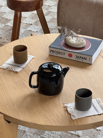 A Serax Passe-Partout Teapot – Black and two ceramic cups on coasters rest on a round wooden table. A copy of Project Japan and a decorative piece with dried leaves complete the minimalist scene.