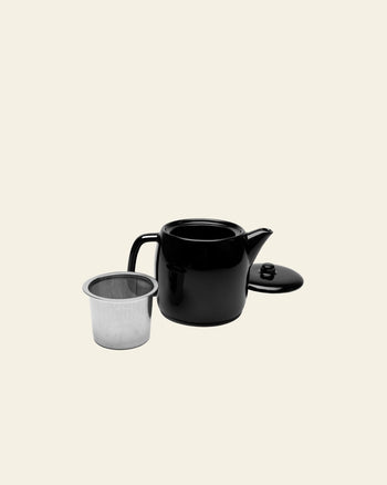 The Serax Passe-Partout Teapot - Black by Serax, with its lid removed, rests beside a metal infuser on a light beige background, highlighting its minimalist design.