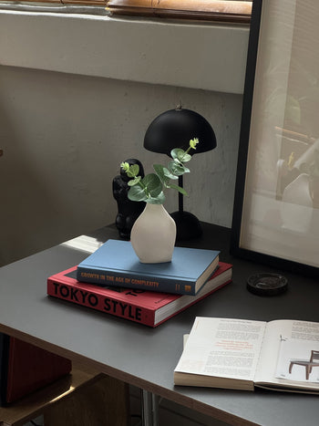 A Serax Perfect Imperfection Flower Vase S N°1 - White by Serax with green leaves sits on a stack of books beside a black lamp and sculpture on a gray desk. Nearby are an open book, a framed picture, and a round dark object.