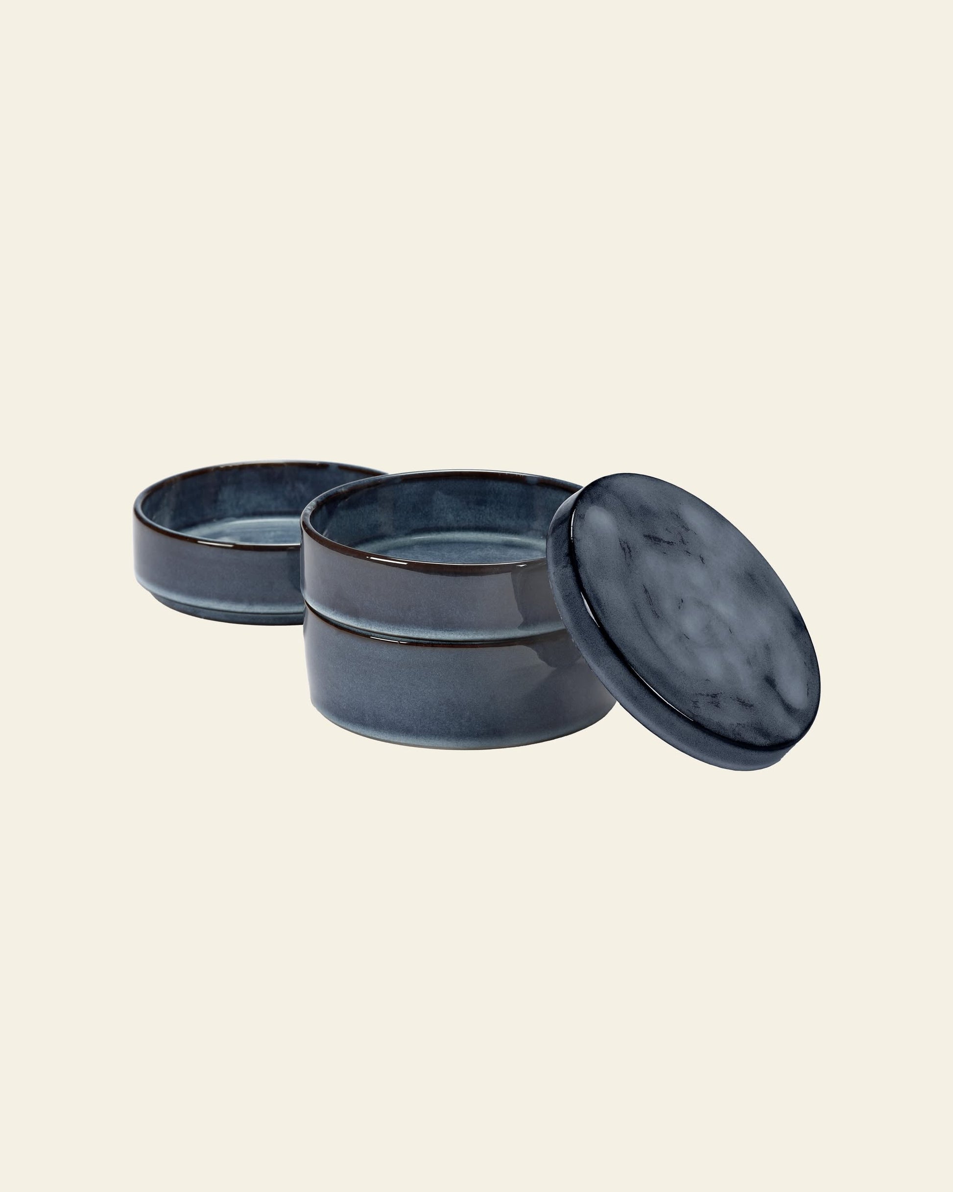 The Serax Pure Stackable Bowl S, Set of 3 in dark blue features glossy, handcrafted stoneware bowls with an earthy finish, stacked with the top lid slightly askew. Set against a plain light background. Brand: Serax.