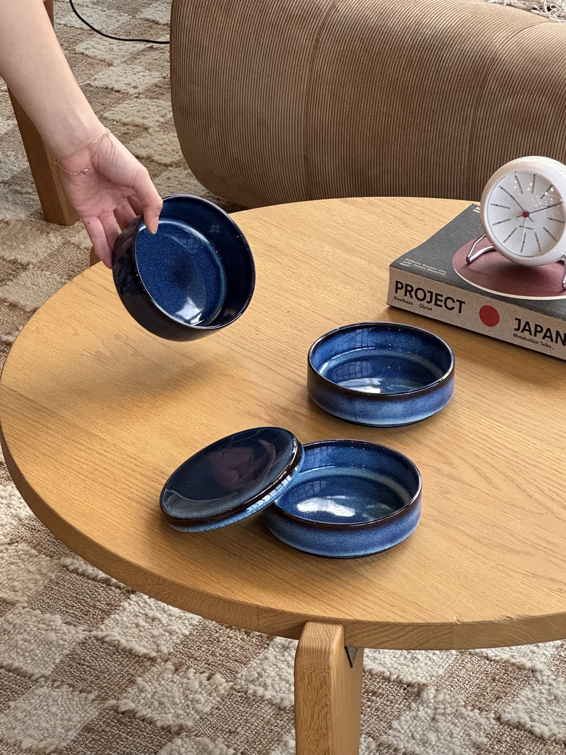 A hand holds a Serax Pure Stackable Bowl S, Set of 3 - Dark Blue above a round table with two more bowls, a clock, and books labeled PROJECT and JAPAN. A beige couch and patterned rug complete this Serax collection scene.