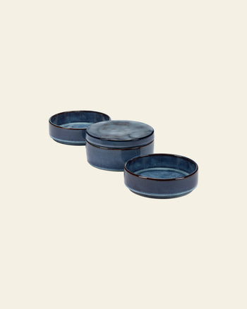 Three Serax Pure Stackable Bowl S dishes in dark blue are arranged in a row on a light beige background; the middle bowl is lidded while the others are open, showcasing their handcrafted stoneware quality.