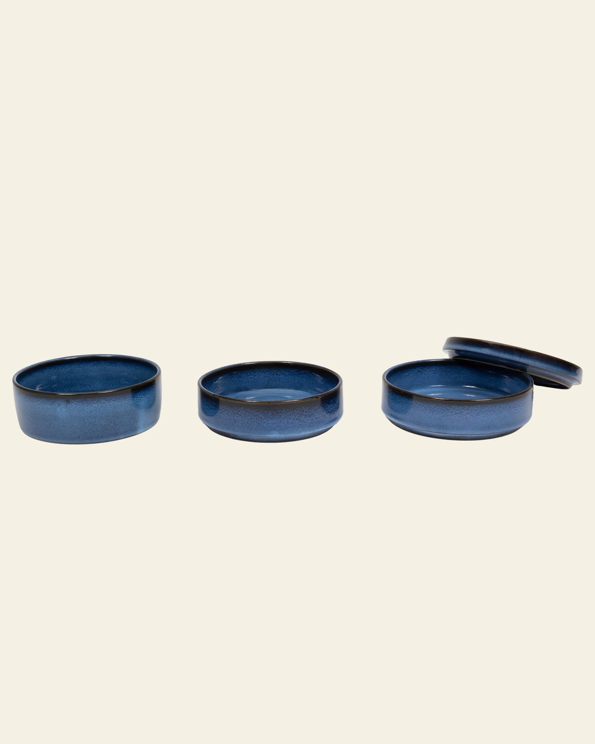 The Serax Pure Stackable Bowl S, Set of 3 - Dark Blue by Serax features three round, handcrafted stoneware bowls. Two are open while one has its matching blue lid placed slightly askew. All are set against a simple light beige background.