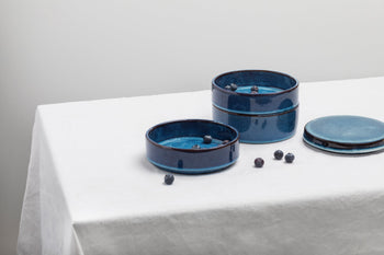 Two Serax Pure Stackable Bowl S dishes, set of 3 in dark blue, sit on a white tablecloth—one open with blueberries inside and one closed with the matching lid, showcasing their earthy glaze and contemporary Serax design.