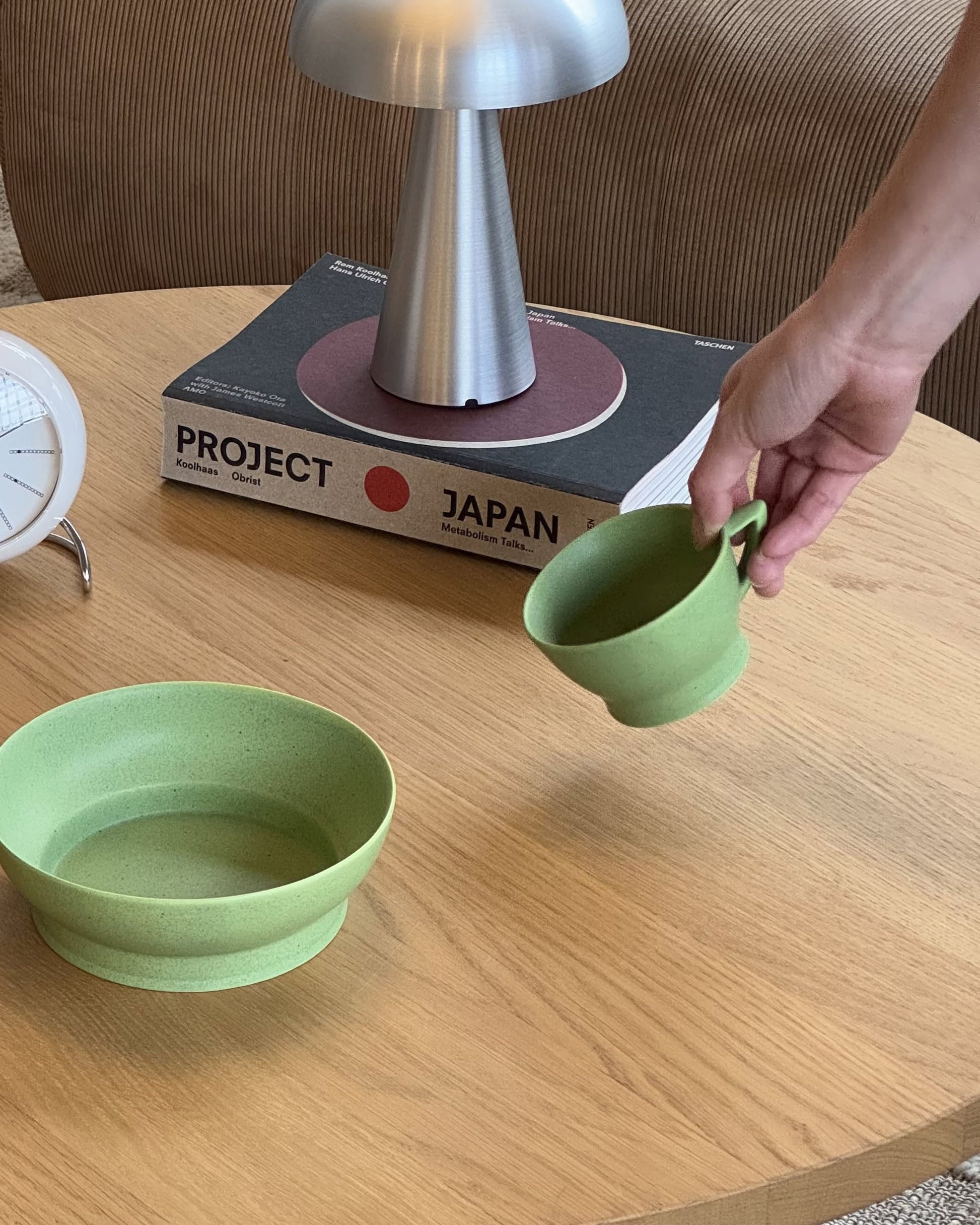 A hand holds a green cup above a wooden table, which also displays the Serax Ra Bowl S - Green by Serax, elegant tableware, a modern lamp, the book Project Japan, and a white clock.