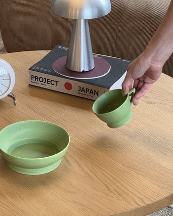 A hand holds a green cup above a wooden table, which also displays the Serax Ra Bowl S - Green by Serax, elegant tableware, a modern lamp, the book Project Japan, and a white clock.