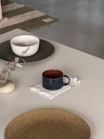 The Serax Terres De Rêves Coffee Cup by Serax sits on a white fringed cloth atop a light table, paired with a beige bowl, brown placemat, and small vase of dried leaves. A textured rug covers the gray floor in the background.