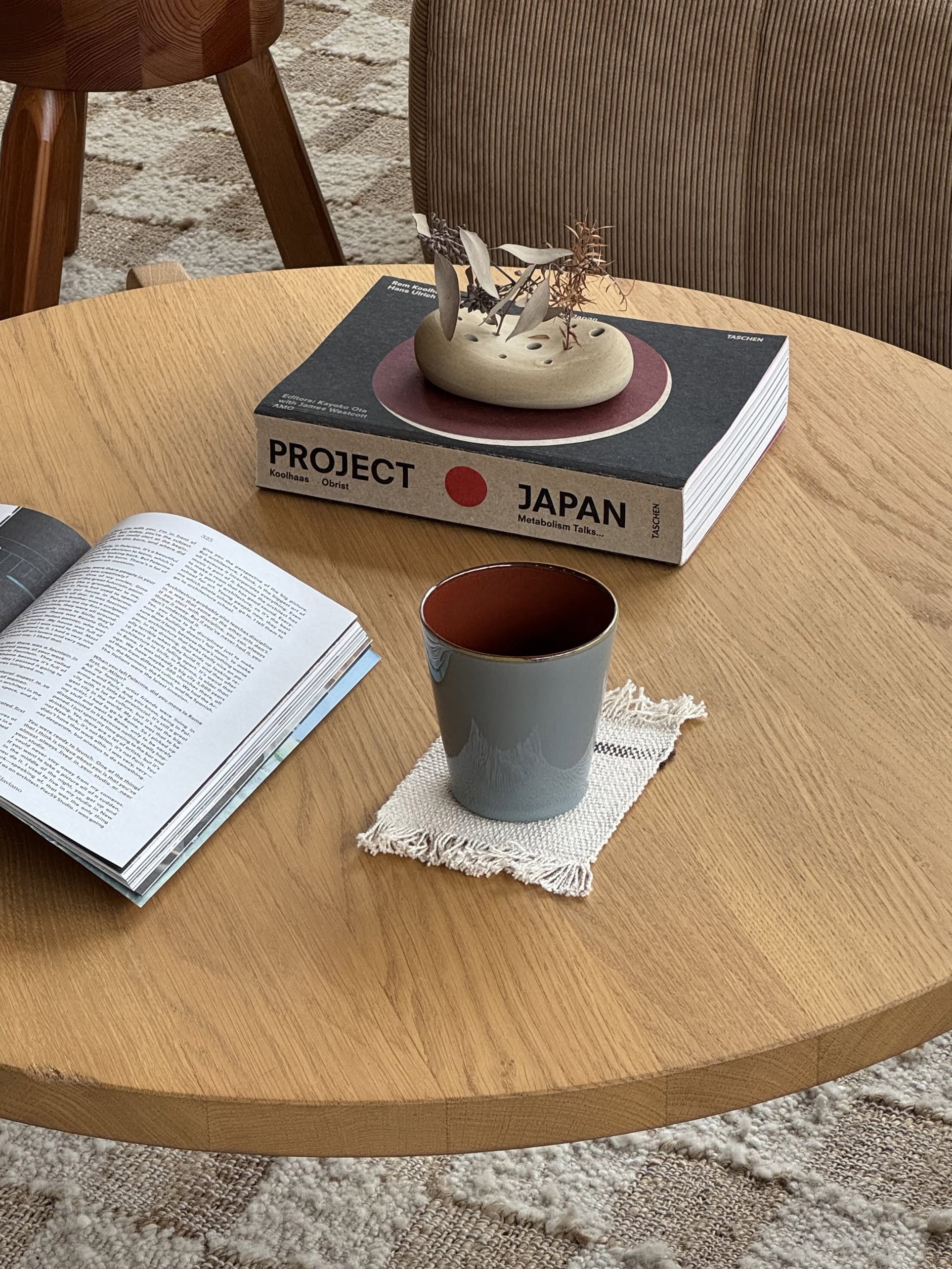 A wooden table holds a Serax Terres De Rêves Conical Cup M in gray with handcrafted glaze on a woven coaster, beside an open magazine and Project Japan topped with a ceramic piece; a chair and patterned rug are in the background.
