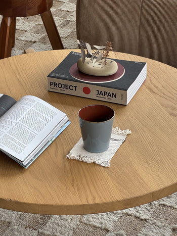A wooden table holds a Serax Terres De Rêves Conical Cup M in gray with handcrafted glaze on a woven coaster, beside an open magazine and Project Japan topped with a ceramic piece; a chair and patterned rug are in the background.