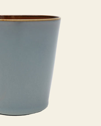 A close-up of the Serax Terres De Rêves Conical Cup M in matte blue with a brown rim, showing its handcrafted glaze. The elegant ceramic cup by Serax is mostly cropped and set against a plain, light background.