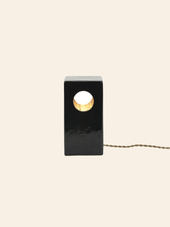 The Serax Terres De Rêves Maxime N°1 Table Lamp in black features a rectangular stoneware base with a central circular hole, emitting warm light. Its geometric design and twisted cord stand out against a plain beige background.