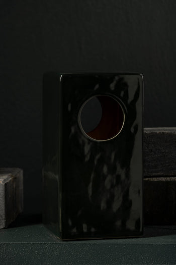 The Serax Terres De Rêves Maxime N°1 Table Lamp in glossy black stoneware features a rectangular shape with a round top hole, standing out against dark bricks and a black wall.