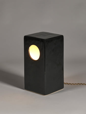 The Serax Terres De Rêves Maxime N°1 Table Lamp in black features a rectangular stoneware base with a round side hole, warm light, and a twisted fabric cord. Its geometric Serax design stands out on a neutral gray surface with a soft shadow.