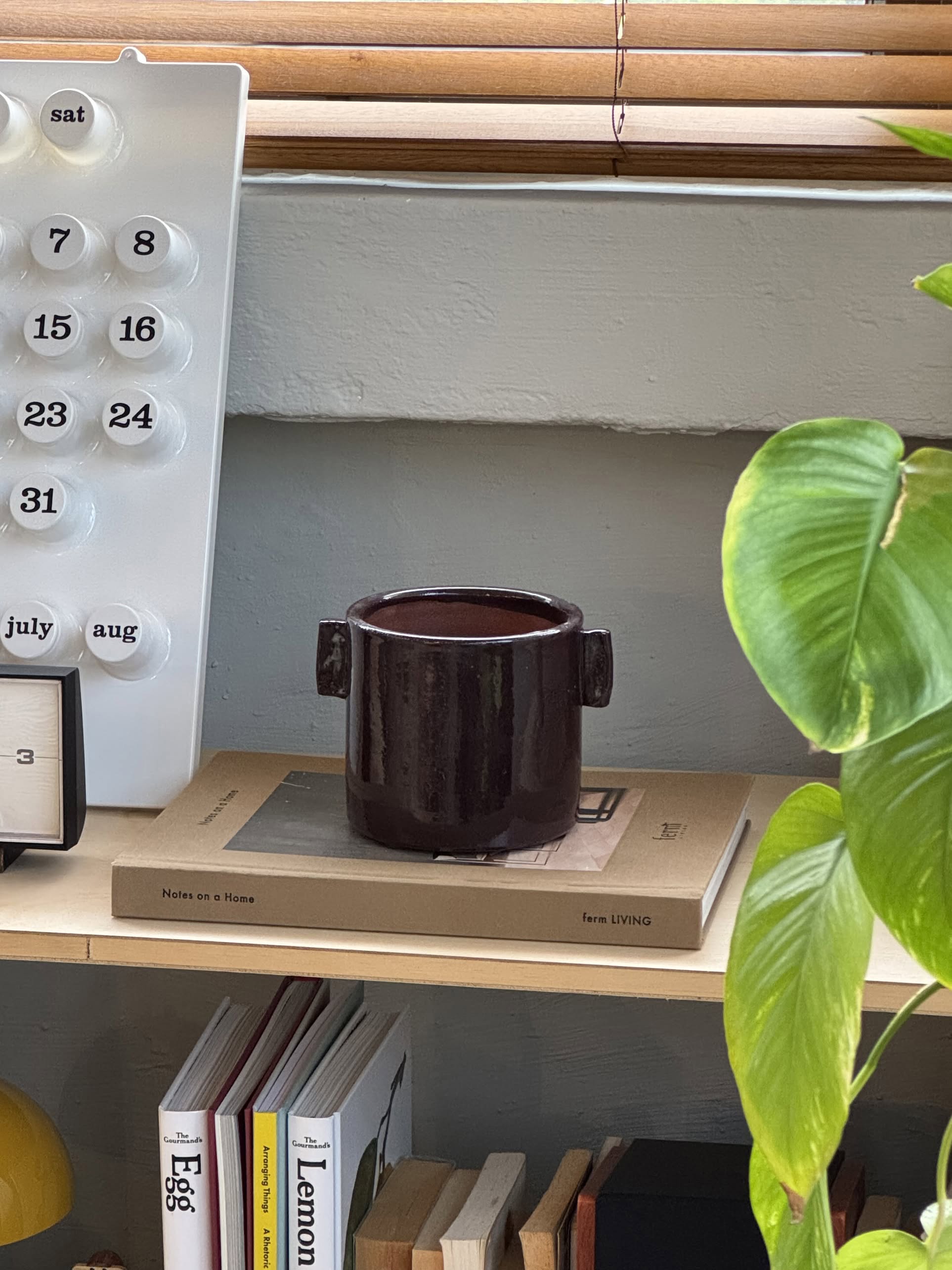 The Serax Urban Jungle Glazed Flower Pot M in Aubergine sits on a beige book atop a wooden shelf, adding charm to your space. A modern calendar and square clock are behind it, with leafy plants and stacked books below.