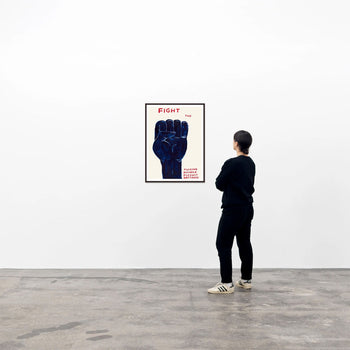 In a minimalist gallery, a person admires the Shrig Shop David Shrigley - Fight (50x70) poster, featuring a large dark blue fist with the word FIGHT in bold red letters at the top.