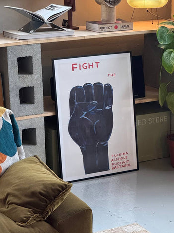 A Shrig Shop David Shrigley - Fight (50x70) poster leans against a shelf in a cozy room, featuring a raised black fist with bold red text and provocative phrases.