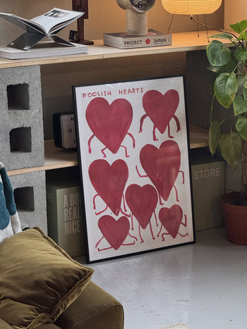 From Shrig Shop, the Foolish Hearts (50x70) framed artwork inspired by David Shrigley shows eight red, heart-shaped figures with arms and legs in a cozy modern room with books and plants.