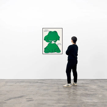 In a gallery, someone admires the Shrig Shop David Shrigley - Frog (Front Of), Frog (Back Of) (60x80) artwork—a large, framed cartoon green frog with FROG above it—displayed on a white wall.
