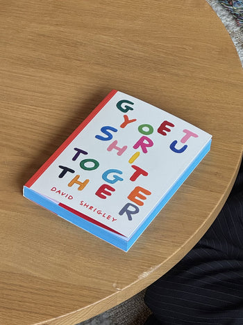 Shrig Shop David Shrigley - Get Your Shit Together, a vibrant humour gift book from Shrig Shop packed with colourful artworks, sits on a wooden table beside someone in pinstripe pants.