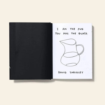 An open Shrig Shop David Shrigley – I Am The Jug You Are The Glass book shows a black blank page on the left and, on the right, a sketch of a jug with the handwritten text “I AM THE JUG YOU ARE THE GLASS” above it.