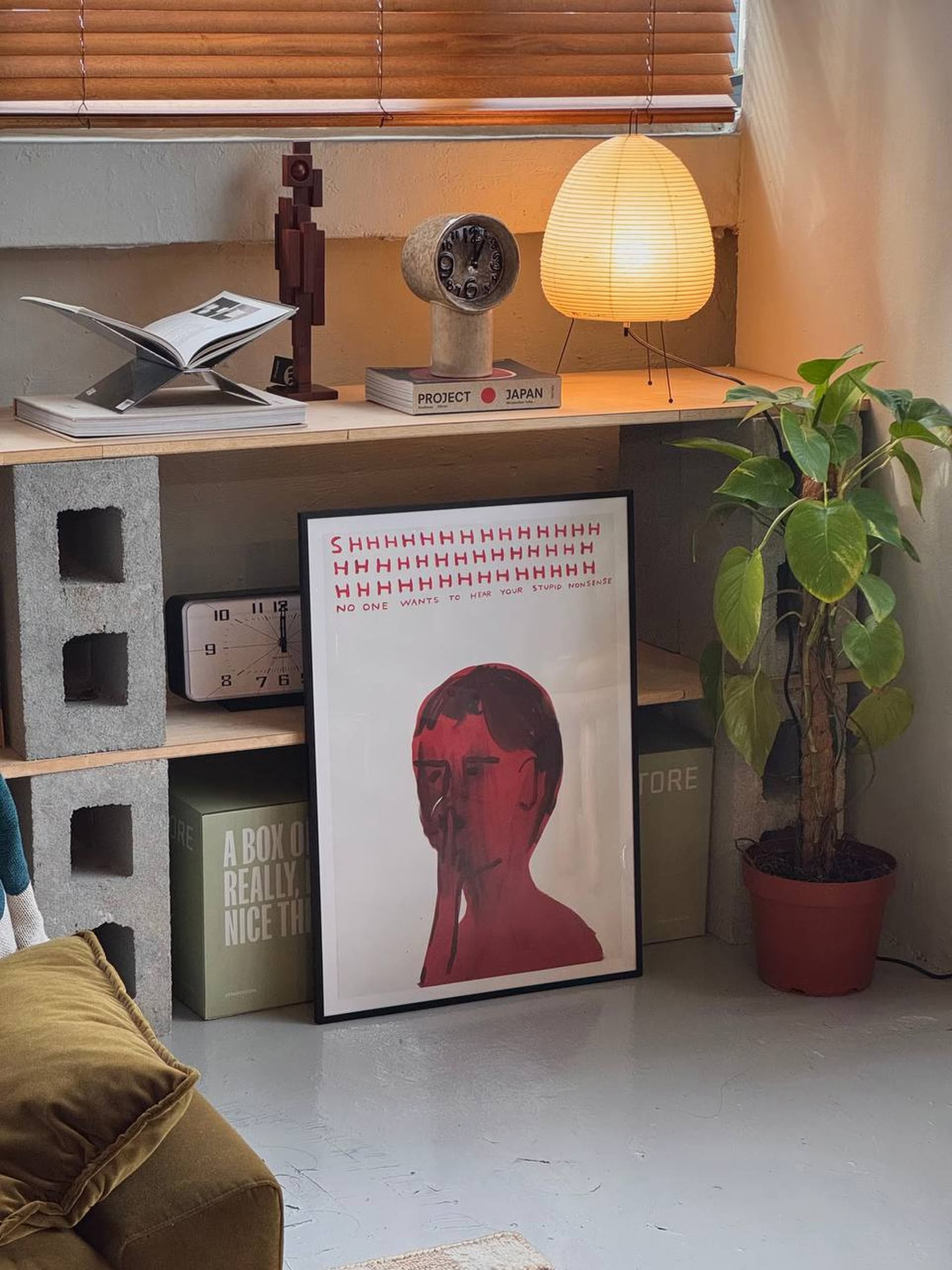 A cozy corner with a wooden shelf, potted plant, and Shrig Shop David Shrigley - Shhh (50x70) artwork. Books, lamp, clock, and sculpture sit on the shelf; box and pillow on the floor—ideal for any Shrig Shop fan or poster lover.