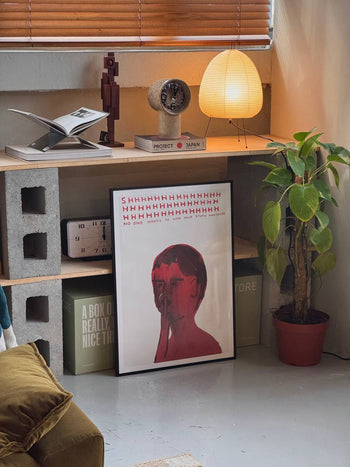 A cozy corner with a wooden shelf, potted plant, and Shrig Shop David Shrigley - Shhh (50x70) artwork. Books, lamp, clock, and sculpture sit on the shelf; box and pillow on the floor—ideal for any Shrig Shop fan or poster lover.