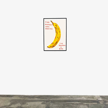 A framed Shrig Shop David Shrigley - The Moment Has Arrived (60x80) print featuring a ripe banana and the text The moment has arrived. The banana is ripe. hangs on a white wall above a gray concrete floor.