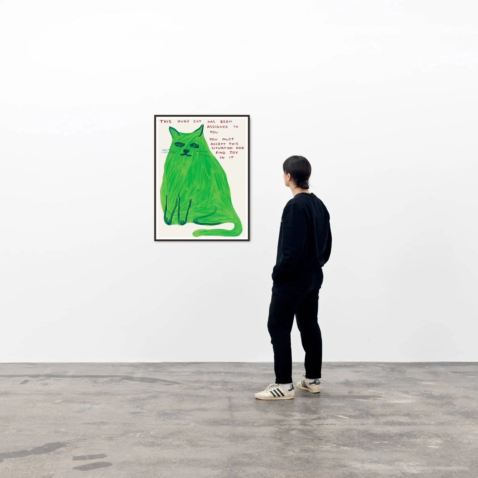 In a minimal white-walled gallery with concrete floors, someone in dark clothes admires the Shrig Shop David Shrigley - This Huge Cat (60x80) poster, featuring a large green cat and handwritten text. Unique art prints stand out in the spacious setting.