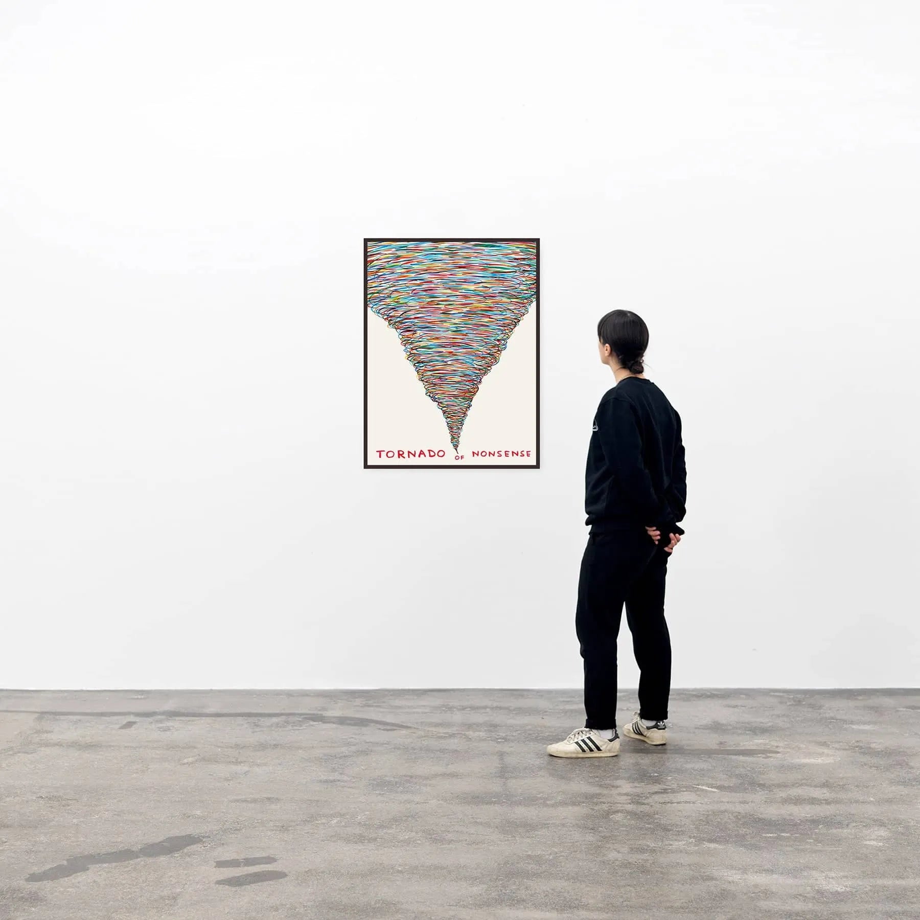 A person in black clothing and white sneakers stands in a gallery, admiring the Shrig Shop David Shrigley - Tornado of Nonsense (60x80) art poster, which features a colorful tornado and bold text at the bottom.